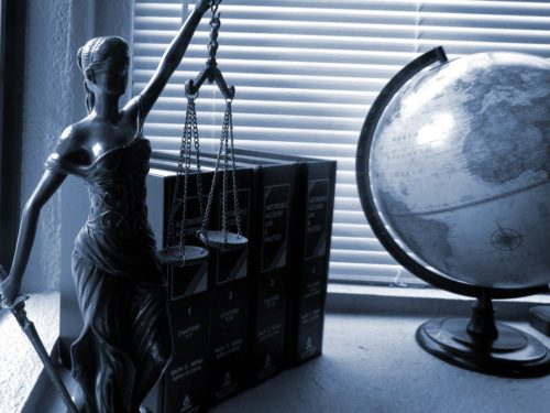 A statue of Lady Justice holding scales stands next to a globe and a row of law books, symbolizing global justice. An experienced attorney's office is suggested by the setting—soft daylight filtering through window blinds, embodying themes of law and expert advocacy.