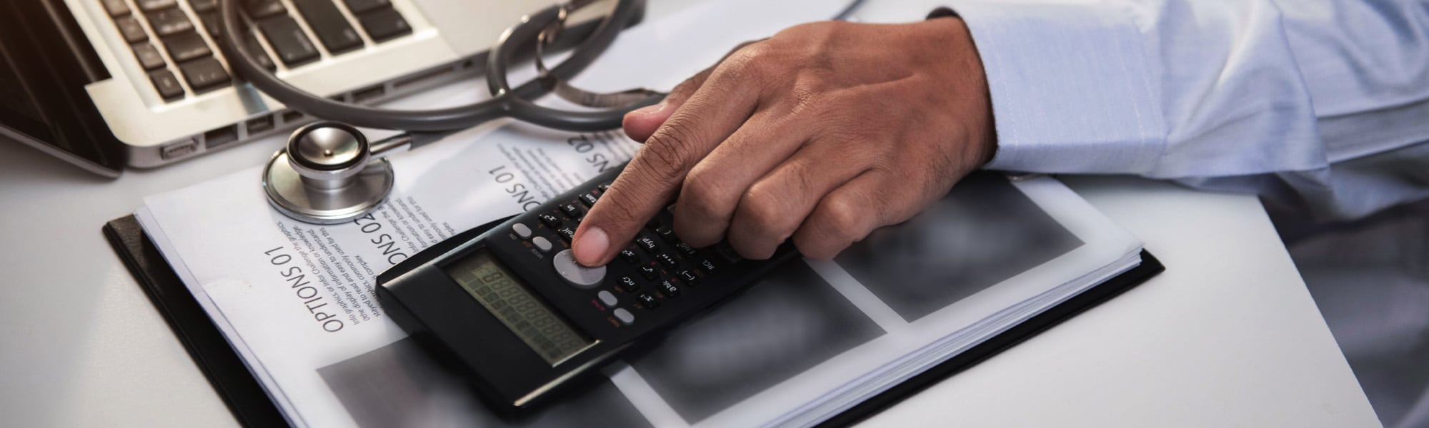 A person is using a calculator, with a stethoscope and laptop nearby. The hand is pressing a button on the calculator, which is placed on a document titled "OPTIONS 01." The scene suggests a combination of medical and financial tasks.