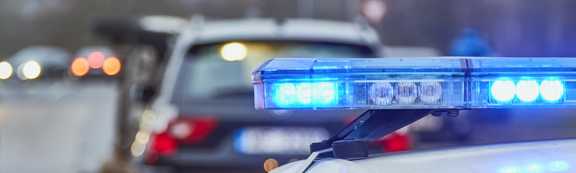 A police car with flashing blue lights is parked beside another vehicle on a road. The scene is slightly blurred, focusing on the emergency lights.