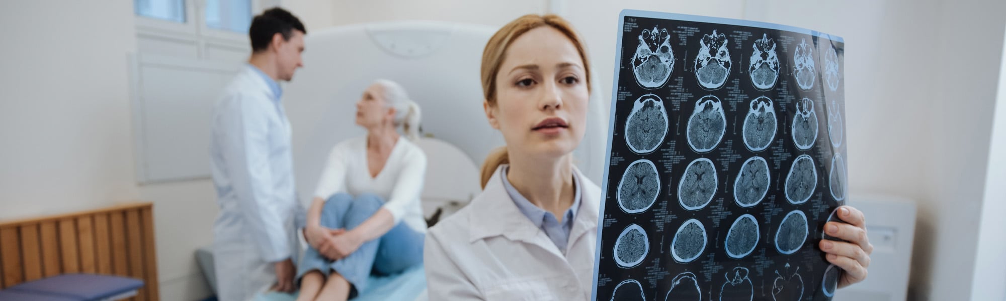 A medical professional examines a brain scan image. In the background, another professional speaks with an elderly patient who is seated and appears attentive. The setting resembles a medical examination room.