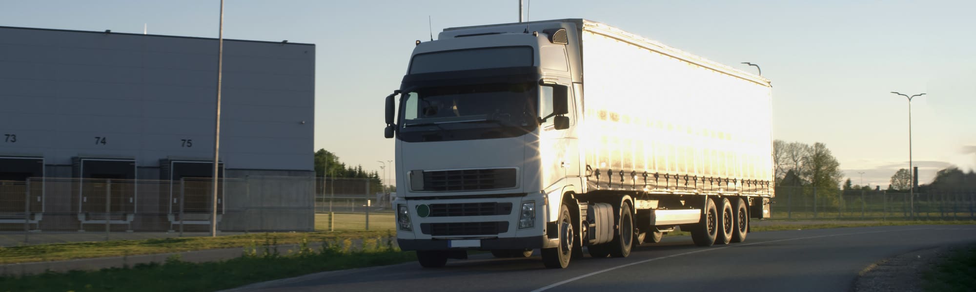 A large white semi-truck with a trailer drives on a paved road at sunset. The background includes a modern industrial building and green grass, with the sky lit by the setting sun.