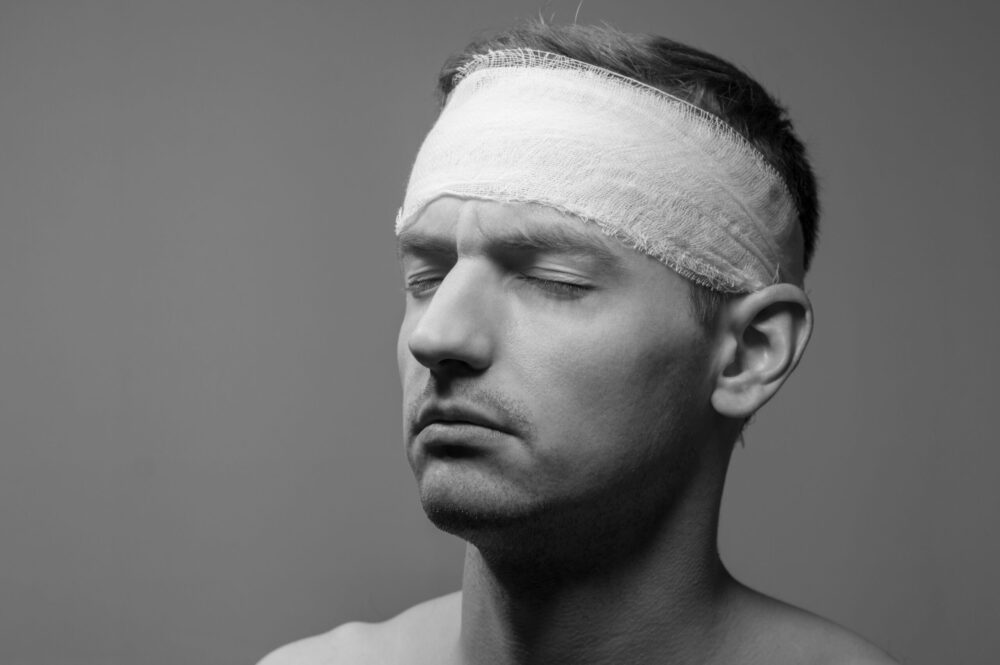 Black and white image of a man with a bandaged head, eyes closed, against a plain background. His expression is calm, possibly reflecting the lingering effects of concussion or chronic fatigue.