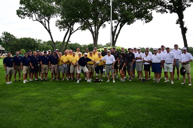 A large group of men stands on a grassy field, posing for a gallery-worthy photo. They are wearing a mix of polo shirts in navy, yellow, and white. Tall trees provide a scenic backdrop under the cloudy sky.