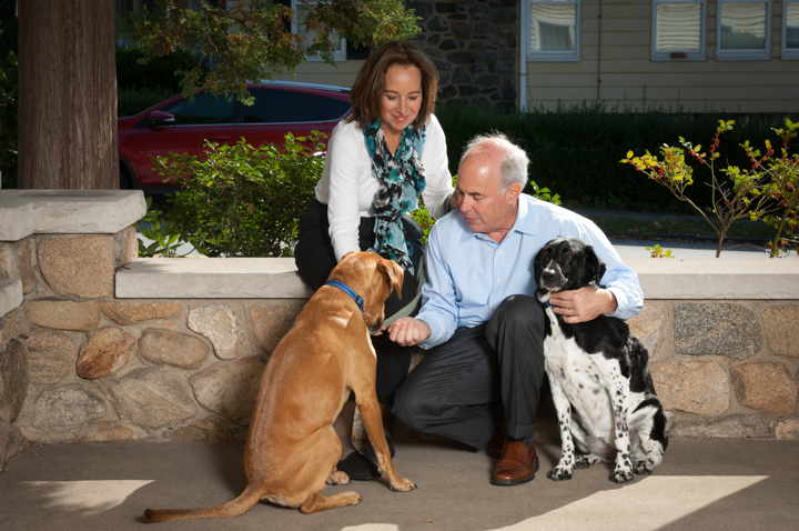 In a charming photo, a man and woman sit on a stone bench with two happy dogs. The man, in a blue shirt, pets a black and white dog while the woman, wearing a white blouse with a blue scarf, smiles warmly at the brown dog. Bushes and a red car form an inviting backdrop.