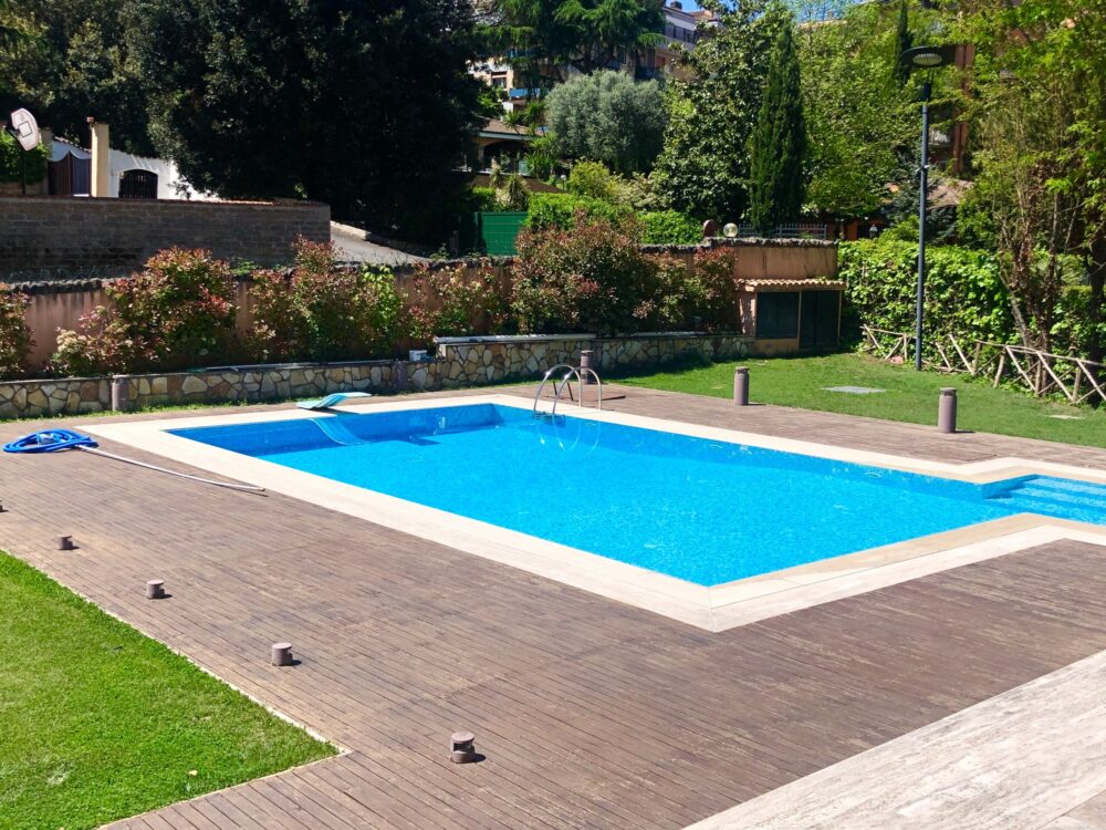 An empty swimming pool, nestled in a Connecticut backyard, rests surrounded by a wooden deck and lush green grass. Trees and bushes line the fence under a clear blue sky while a cleaning net lies on the deck, ready for use.
