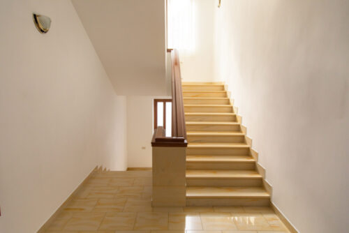 A well-lit indoor stairway with wooden railings and tiled steps leads to a higher floor. The neutral-toned walls and flooring create a bright, airy space, enhanced by natural light from a window above, ensuring safety and minimizing the risk of accidents.