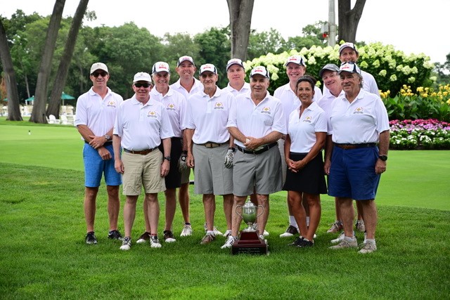 A group of eleven people wearing white shirts and hats pose on a golf course, creating a perfect moment for any photo gallery. They stand around a trophy on the grass, with trees and flowerbeds enhancing the picturesque backdrop.