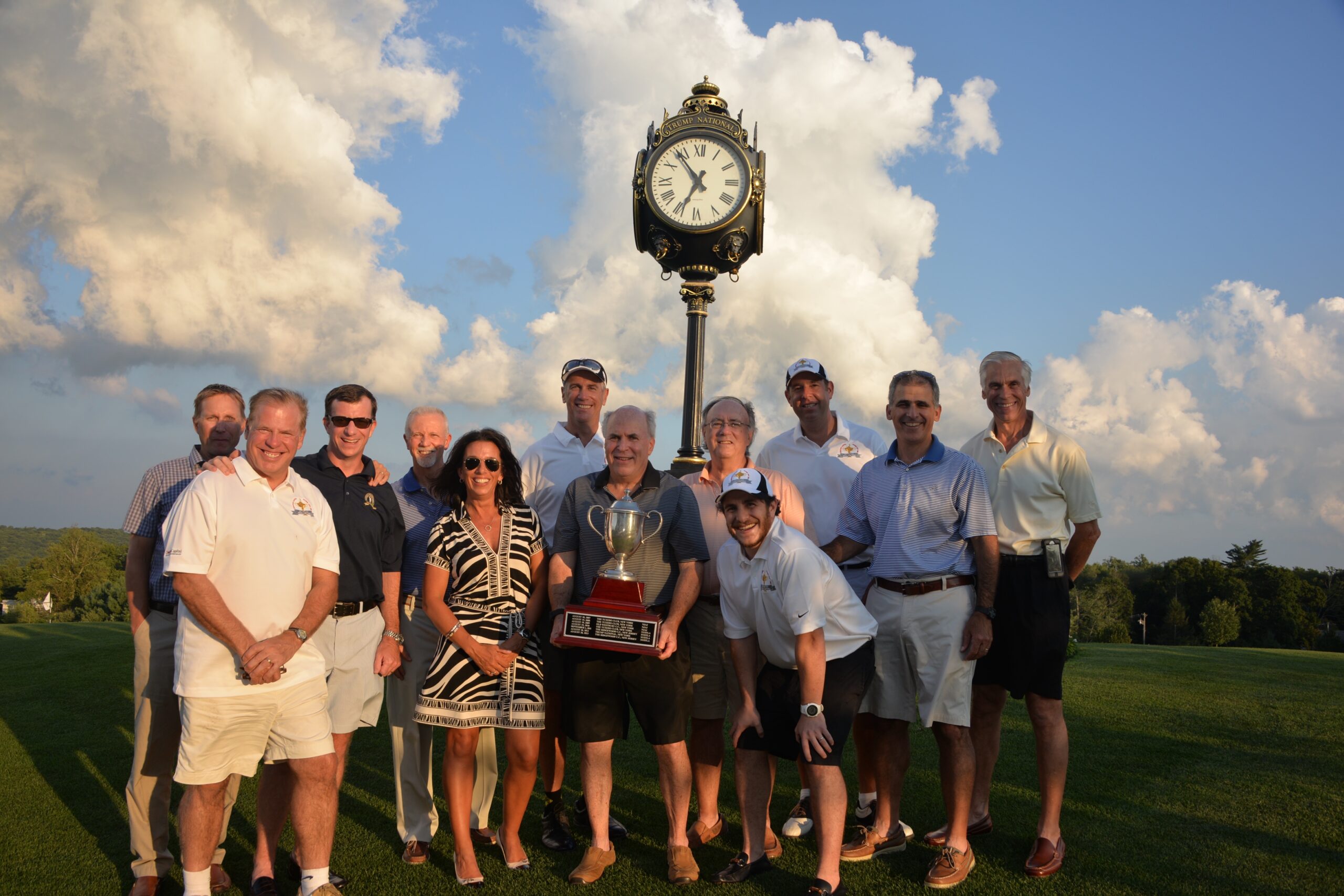 A photo captures a group of people standing together on a golf course, smiling with a trophy in front. Behind them, a vintage clock tower stands proudly under the blue sky with clouds, creating a perfect gallery-worthy moment.