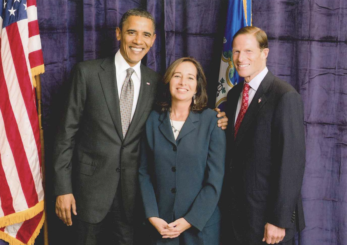 In this elegant photo, three people in formal attire stand together in front of a purple curtain, with U.S. and Connecticut flags visible. The two men wear suits and ties, and the woman in the center is wearing a dark suit. They are smiling at the camera, creating a striking gallery-like composition.