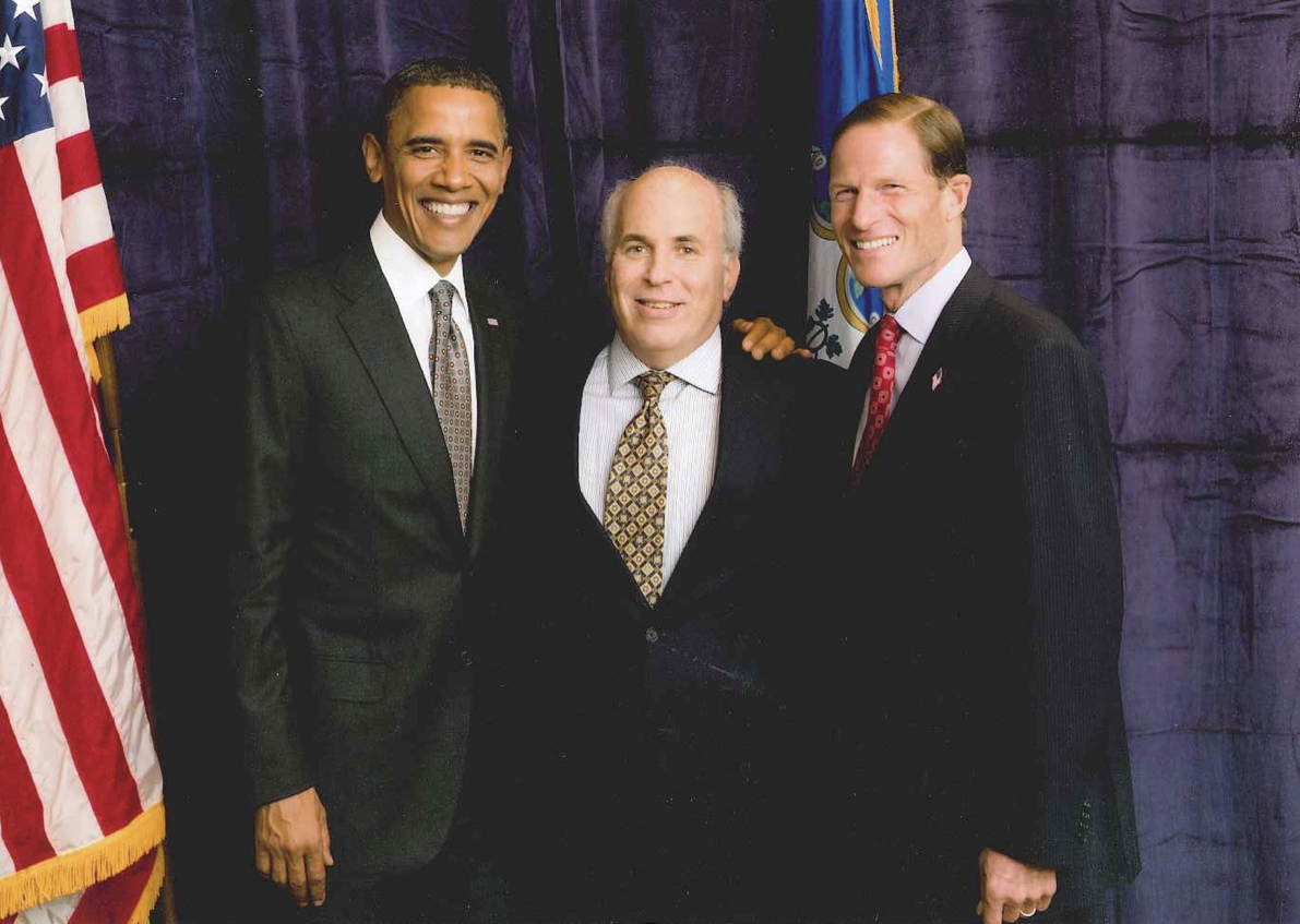 Three men in business attire stand together, smiling, in front of a dark curtain with an American flag and another flag partially visible in the background, capturing a moment perfect for any professional photo gallery.