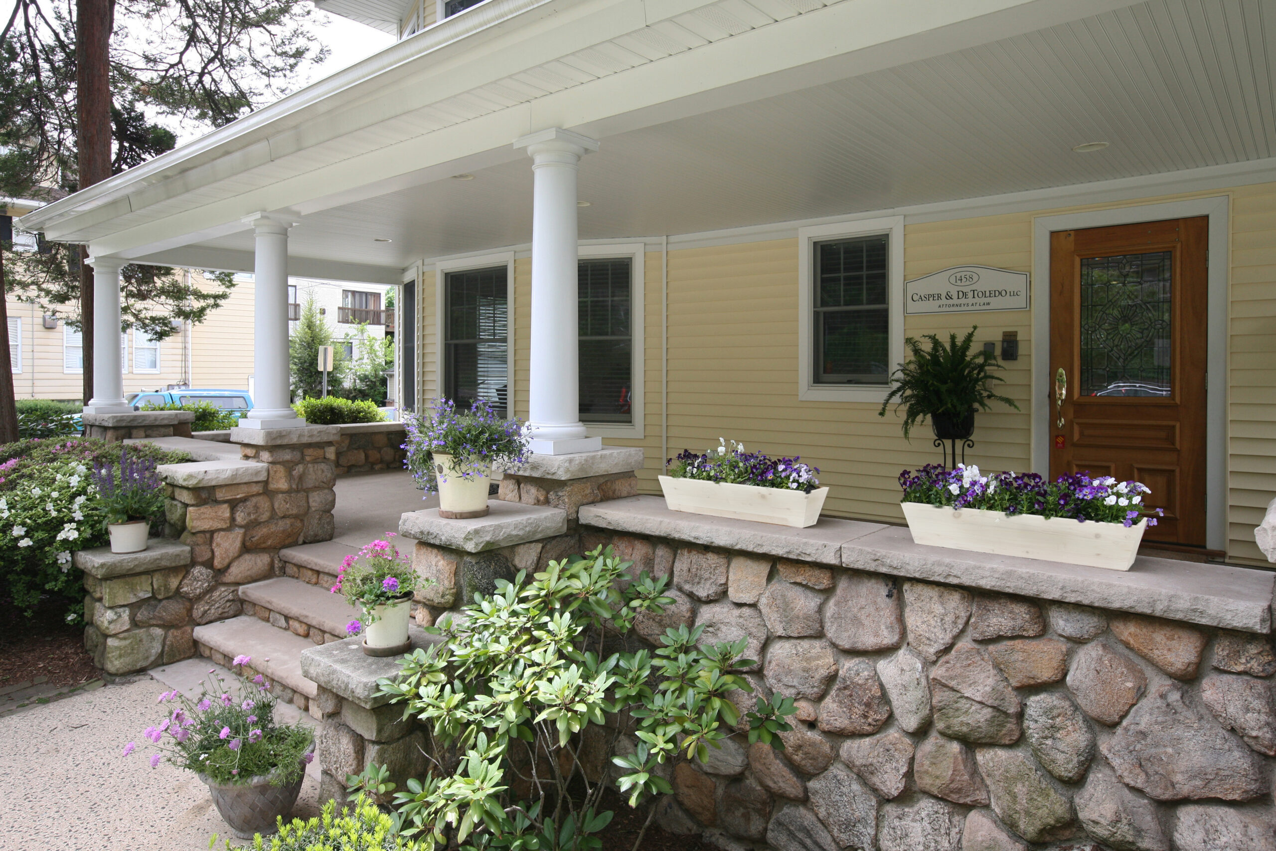A charming house with a porch featuring white columns, stone steps, and planters filled with purple and white flowers. A wooden door with a glass pane is on the right, and the siding of the house is painted yellow.