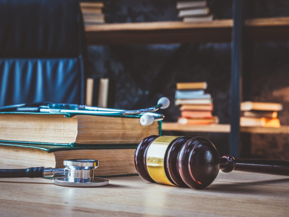 A wooden gavel rests on a table beside a stack of thick books and a stethoscope, hinting at the intricate dance between law and medicine. Shelves filled with more volumes loom in the background, evoking thoughts of negligence cases in Connecticut's legal landscape.