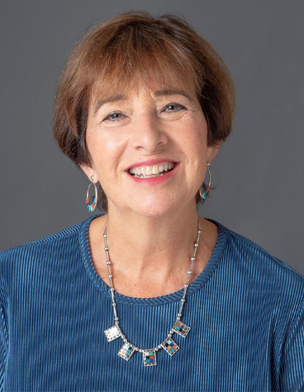 A smiling woman, reminiscent of a Victoria de Toledo muse, has short brown hair. She's clad in a blue top, hoop earrings, and a necklace with square pendants. The backdrop is gray.