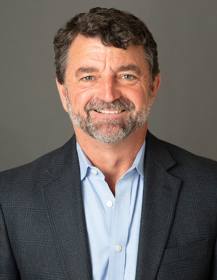 Daniel T. Saltus, a man with curly dark hair and a beard, smiles at the camera. He is wearing a blue shirt and a dark blazer against a plain gray background.