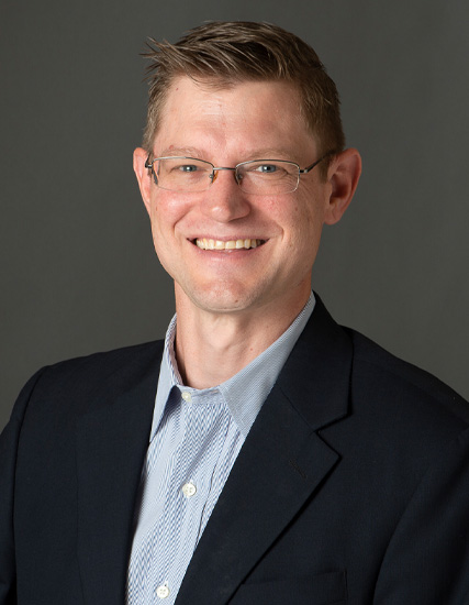 Daniel T. Saltus, a man with short brown hair and glasses, smiles at the camera. He is wearing a dark suit jacket over a light blue collared shirt against a plain gray background.