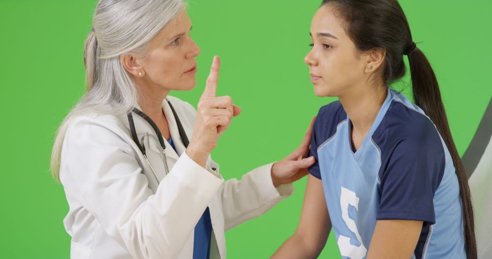 A doctor with gray hair and a stethoscope gestures with a finger, discussing PINK Concussions with a young woman in a sports jersey. The young woman looks attentive, absorbing the important information. The background is a solid green color.