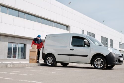 A delivery driver wearing a blue hat and red jacket is carefully unloading cardboard boxes from a white van parked outside a large, modern industrial building.