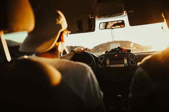 A person wearing a cap drives with an edge during sunset. The car's interior is visible, sunlight streaming in through the windshield, casting a warm glow that contrasts with the aggressive driving style.