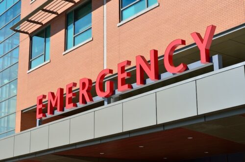 A red "EMERGENCY" sign adorns the hospital's exterior, where brick meets glass and windows soar above, guiding those from an auto accident to aid. The sign prominently marks the entrance, a beacon for when medical bills and urgent care collide in critical moments.