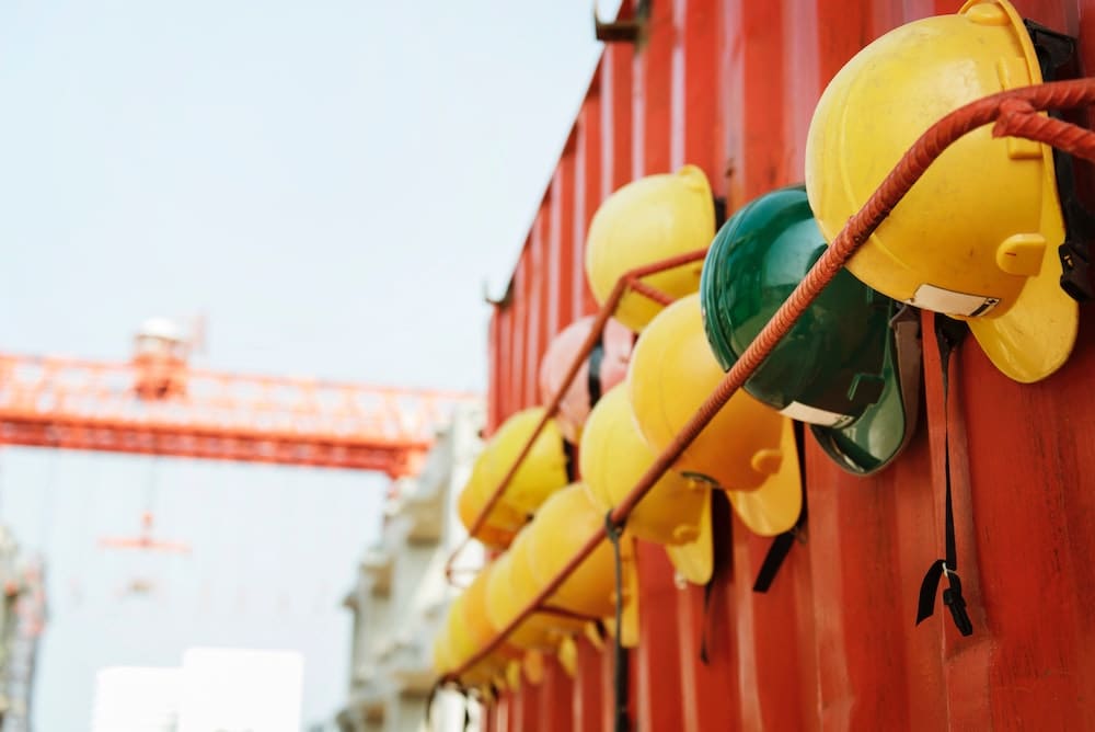 A row of yellow and green construction helmets hang on a red container wall, resembling the caution necessary in avoiding mistakes akin to those in a car accident, with a blurred construction crane visible in the background against a clear sky.