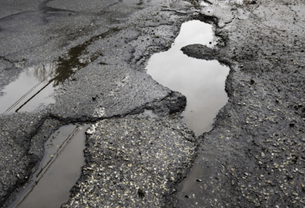 A close-up of a damaged road with large potholes filled with water, possibly leading to car accidents. The surrounding asphalt is cracked and uneven, reflecting the gray sky.