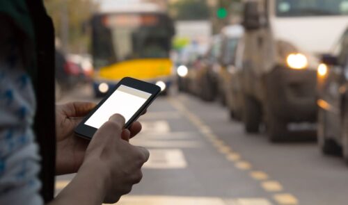 A person uses a smartphone while standing on a bustling urban street, unaware of the potential risks. Cars and a yellow bus navigate the background, with blurred traffic emphasizing motion, highlighting how easily a small mistake could lead to a car accident in such chaos.