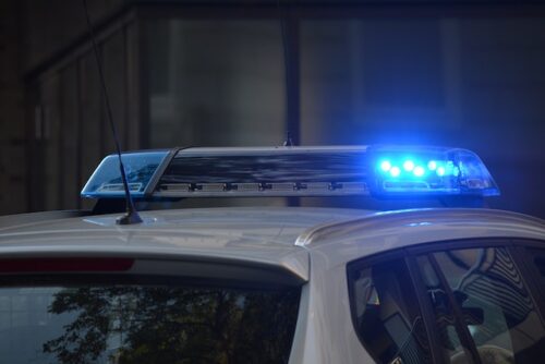 Close-up of a police car's rooftop with flashing blue lights, hinting at a recent accident. The background is blurred, highlighting the vehicle's light bar in an urban setting.