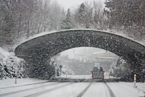 A car cautiously drives on a snow-covered road under a stone bridge. Heavy snowfall obscures the background, with trees lining either side, hinting at the potential for winter auto accidents. The scene captures a typical winter day with a serene, wintry ambiance.