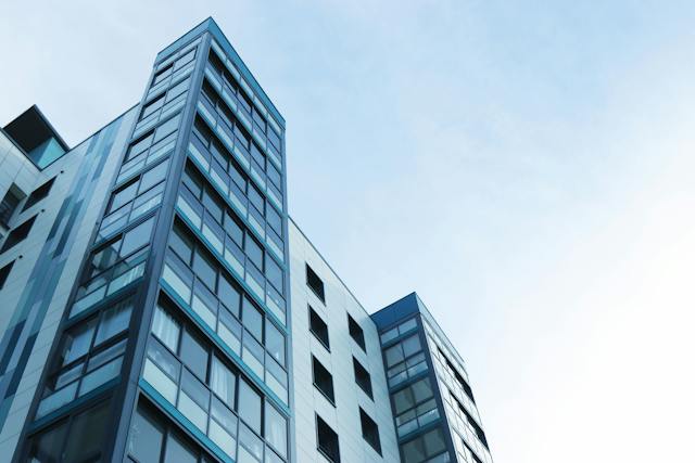 Low-angle view of modern high-rise buildings with glass windows against a clear blue sky. These sleek, contemporary structures rise proudly, each story overseen by vigilant landlords ensuring safety and elegance in every detail.