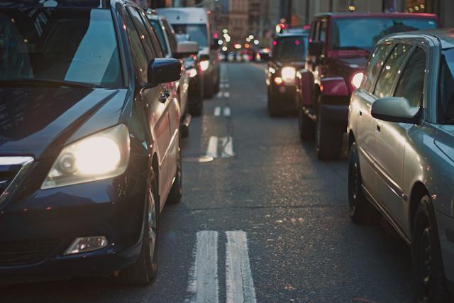 Heavy traffic with cars lined up on a city street during dusk hints at a multi-car accident. Headlights illuminate the road, casting long shadows as blurred buildings stand in the background.