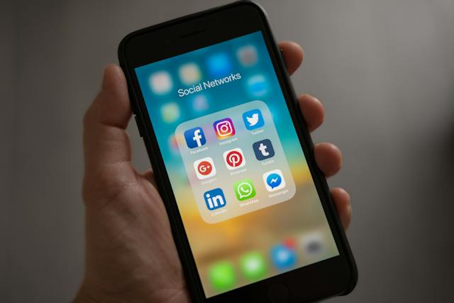 A hand holds a smartphone displaying a folder titled "Social Networks" with popular social media apps: Facebook, Instagram, Twitter, Google+, Pinterest, Tumblr, LinkedIn, and WhatsApp. The background is blurred.