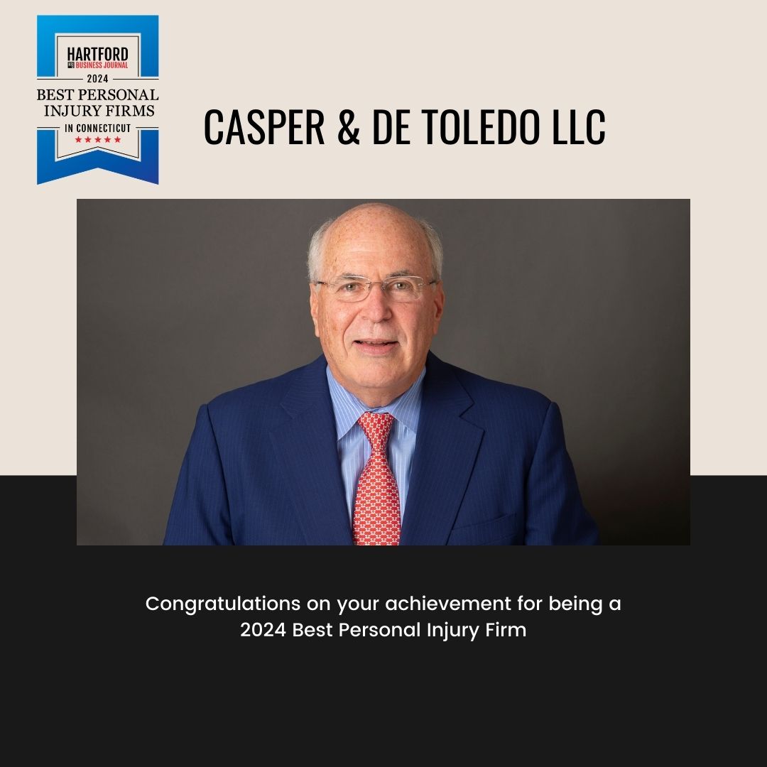 An older man in a blue suit and red tie stands before a gray background. A badge labeled "Hartford 2024 Best Personal Injury Firms" graces the upper left, with text applauding Casper & De Toledo LLC for their recognition. Featured in Hartford Business Journal as a 2024 Best Personal Injury Firm.