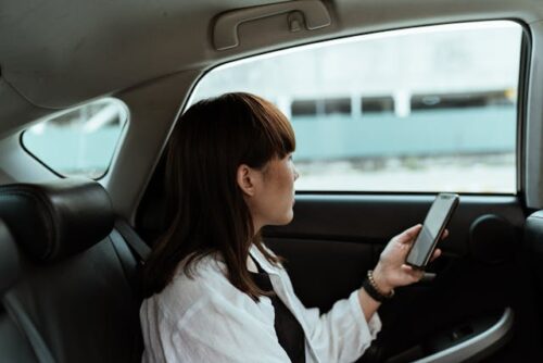 A passenger with shoulder-length hair sits in the backseat of a car, holding a smartphone and gazing out the window. Clad in a white shirt, they seem lost in thought as the world outside blurs by.