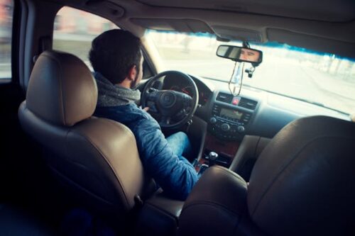 A person with dark hair, wrapped in a blue jacket and scarf, is driving a car, possibly an uninsured motorist. The photo captures the scene from the back seat, showcasing their hands on the steering wheel and the road unfurling through the windshield. The interior displays hues of brown and gray.