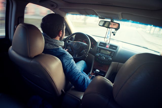 A person with dark hair, wrapped in a blue jacket and scarf, is driving a car, possibly an uninsured motorist. The photo captures the scene from the back seat, showcasing their hands on the steering wheel and the road unfurling through the windshield. The interior displays hues of brown and gray.