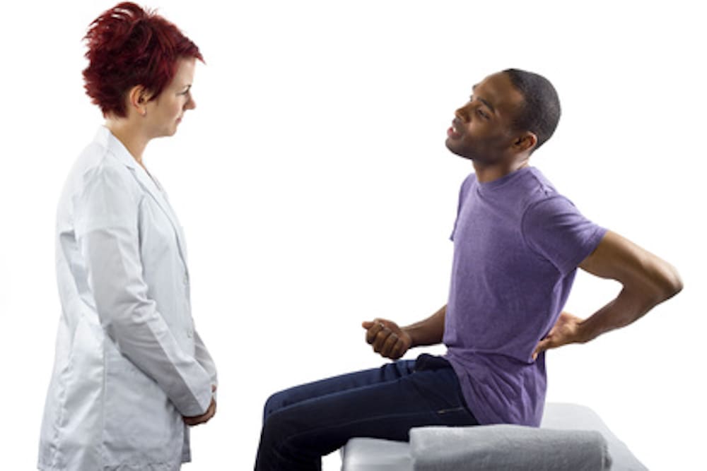 A doctor in a white coat is attentively listening to a patient sitting on an examination table. The patient, discussing his back pain and possible pre-existing conditions, holds his lower back as they engage in a focused conversation.