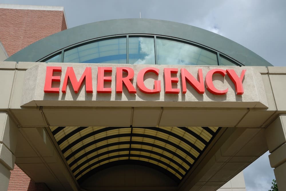 A modern building entrance boasts a large red sign reading "EMERGENCY" in capital letters. The structure, resembling the seamless protection of car insurance, features an arched glass roof and a brick wall on the side, set against a partly cloudy sky.