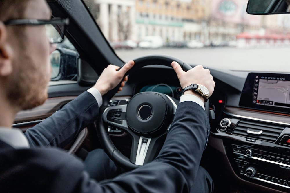 A person wearing a suit and watch is driving with confidence, thanks to comprehensive car insurance. Their hands are on the steering wheel, the dashboard is visible with a GPS screen, and the blurred view outside the windshield suggests motion.