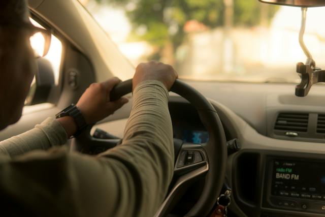 A person is cautiously driving a car with both hands on the steering wheel, mindful of avoiding a rear-end collision. The car's interior, including the dashboard and radio, is visible. Sunlight softly filters through the windows, casting gentle shadows on the driver's focused expression.