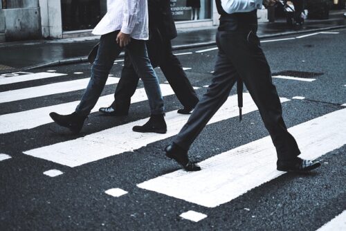 Three people walk cautiously across a zebra crossing on a city street, their feet showcasing diverse styles of footwear like boots and formal shoes. The wet pavement hints at recent rain, reminding pedestrians to tread carefully to avoid any fault or accidents.