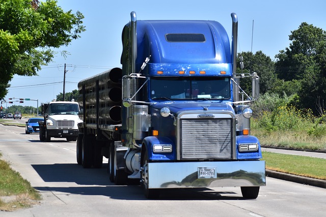 A large blue semi-truck with a trailer gracefully cruises down a multi-lane road, surrounded by cars and lush greenery lining the way. The clear, blue sky creates an idyllic backdrop as the truck moves safely, steering clear of any potential trucking accidents.