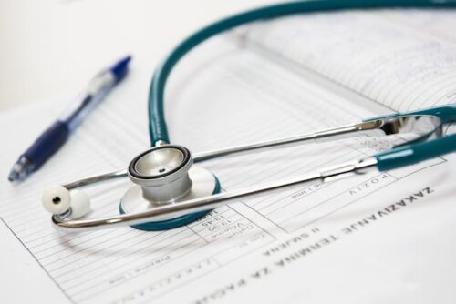 A stethoscope rests on an open medical chart with handwritten notes, perhaps detailing injuries from a car accident. A blue pen lies beside the chart, suggesting a healthcare setting where treatment and medical bills are addressed.