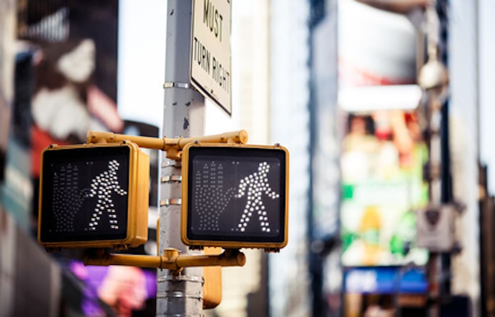 Two pedestrian walk signals show white walking figures against a blurred urban background, emphasizing the importance of safety to prevent pedestrian accidents. The signals are mounted on a pole with a partial "MUST TURN" sign visible above them.