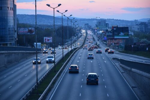 A city highway at dusk features a steady traffic flow, with cars driving in both directions on the multi-lane road. Streetlights cast a glow as buildings and illuminated billboards line the road, creating a vibrant scene as the sky transitions from day to night.