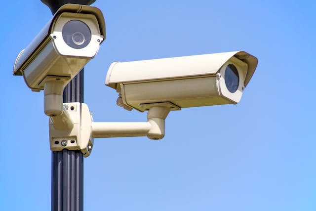 Two white surveillance cameras, poised to capture detailed footage, are mounted on a pole against a clear blue sky.