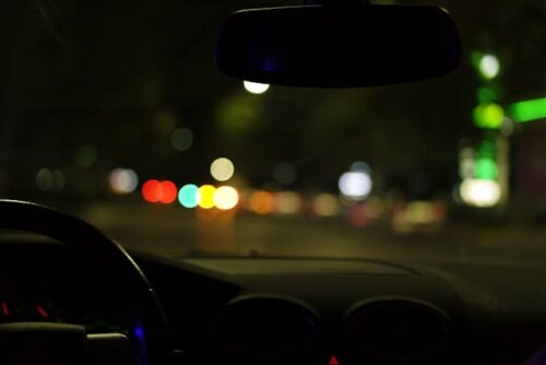 View from inside a car at night. The dashboard and steering wheel are visible in silhouette, capturing the scene before a potential DUI incident. Outside, the streetlights and signs are blurred, creating colorful, circular bokeh effects against the dark background.