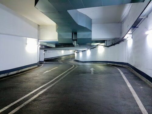 A dimly lit underground parking garage with white walls and a winding, narrow two-lane road. The lanes are separated by dashed white lines, while overhead pipes trail the ceiling, raising questions about liability. Arrows on the road guide drivers through safely to prevent parking garage injuries.