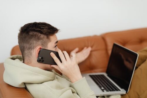 A person in a light-colored hoodie sits on a brown couch, phone to ear and gesturing, likely discussing timely reporting with insurance companies. Meanwhile, a laptop rests open on their lap, perhaps detailing recent car accident claims.