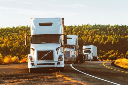 Three semi-trucks, compliant with regulations, drive along a curving highway bordered by trees and greenery under a clear sky, with the sunlight casting warm tones on the scene.